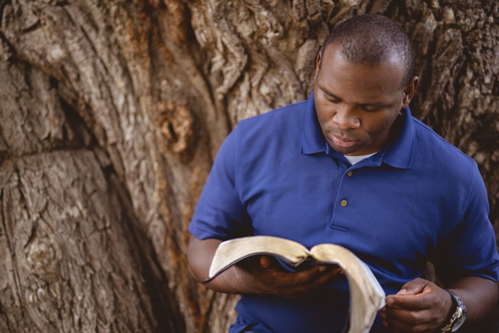 A closeup of an African-American man reading a bible with a tree on the blurry background